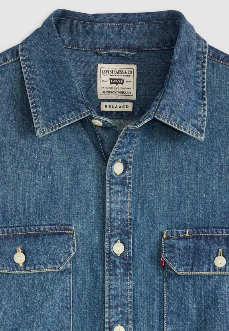 Camicia in denim blu medio con vestibilità comoda, caratterizzata da colletto appuntito, due tasche sul petto, bottoni bianchi e dettagli di cuciture arancioni.