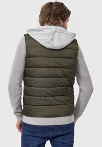 Olivengrøn polstret vest med hætte, parret med en lys grå striktrøje, har vertikal quiltning og ribbede manchetter ved kanten.