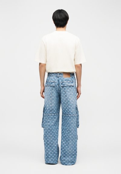 Personne aux cheveux courts noirs portant une chemise unie à manches courtes couleur écrue et un jean large bleu à motifs avec poches latérales, debout de dos.