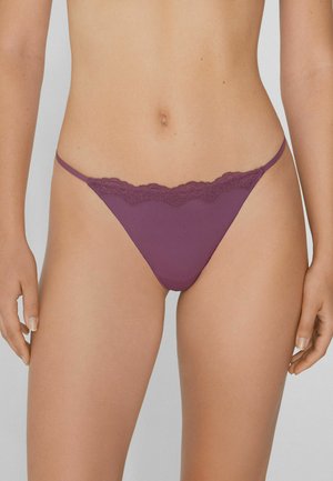 Femme portant des sous-vêtements en bikini avec un ourlet en dentelle violette, se tenant avec les mains détendues sur les côtés devant un fond neutre.