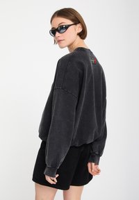 Joven mujer con gafas de sol negras, una sudadera gris oscuro holgada con un pequeño bordado de rosa roja en la parte trasera y pantalones cortos negros, mirando por encima del hombro.