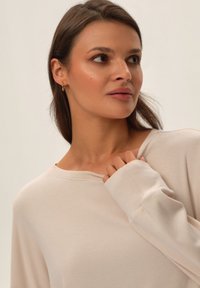 Sweter z długim rękawem w kolorze kremowym, z okrągłym dekoltem. Miękka tkanina o gładkiej strukturze. Modelka pokazuje sweter trzymany na ramieniu.