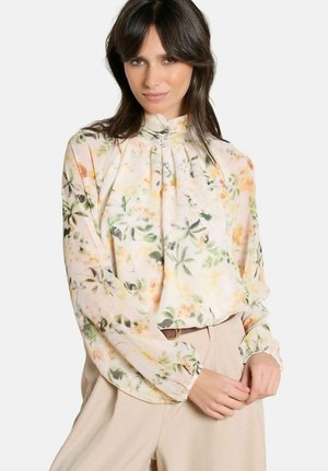 Femme portant un chemisier à manches longues et col haut orné de fleurs aux couleurs pastel douces, et un pantalon beige taille haute, les mains délicatement jointes.