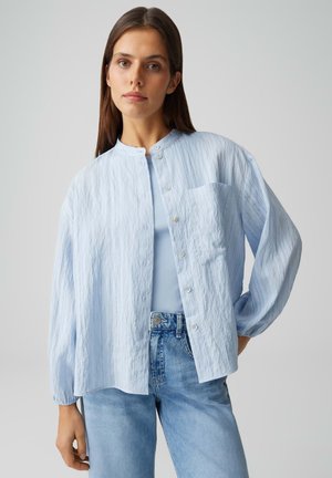 Femme portant une chemise boutonnée à texture bleu clair sur un débardeur bleu, associée à un jean taille haute bleu clair, debout devant un fond uni.