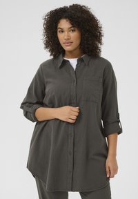 Chemise légère gris foncé à manches longues retroussées aux coudes, avec poche avant et ourlet arrondi. Texture lisse, design décontracté.