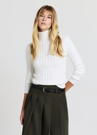 Pull à col roulé blanc à côtes avec un motif en maille texturée, assorti à un pantalon plissé taille haute vert foncé et une ceinture noire.
