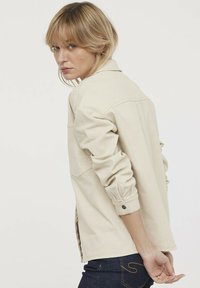 Camisa chaqueta de manga larga en beige, hecha de algodón, con puños abotonados y aberturas laterales. El diseño incluye un yugo trasero y bolsillos.