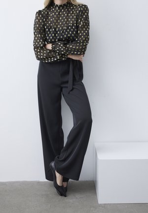 Schwarze transparente Bluse mit goldenen ovalen Mustern, langen Ärmeln und hohem Kragen; kombiniert mit hochtaillierter schwarzer weiter Hose und schwarzen Absätzen.