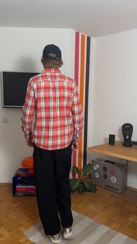 Sarkans un balts pleda krekls, melnas bikses un sudraba kedas. Fonā ir TV, krāsainas sienas svītras un zaļa auga.