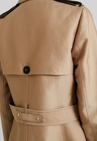 Beżowy płaszcz trencz o prostym designie, z tylnym guzikiem i paskiem w talii. Gładka tekstura, bez widocznych wzorów.