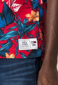 Czerwona tropikalna koszula z niebieskimi i pomarańczowymi wzorami kwiatowymi. Posiada białą metkę z logo "Tommy Jeans" i napisem "Nowy Jork".