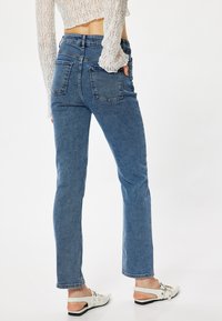 Jeans de mezclilla azul de tiro alto y pierna recta con un acabado desgastado, que cuentan con dos bolsillos traseros y un dobladillo limpio. Llevados con zapatos de tacón abierto blancos.