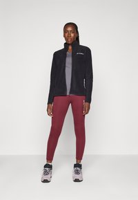 Giacca in pile con zip nera, maglione grigio, leggings bordeaux e scarpe atletiche grigie e blu. La giacca presenta un colletto alto e un logo.