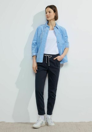 Camicia di jeans azzurro chiaro, t-shirt bianca, pantaloni jogger blu navy con cordino, sneakers bianche. Texture lisce, vestibilità casual, posa in piedi contro un muro.