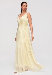 Femme debout portant une robe fluide jaune pâle sans manches avec un profond décolleté en V et un détail froncé à la taille sur un fond clair uni.
