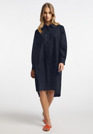 Navyblauwe oversized button-up jurk met lange mouwen, voorzien van een kraag en een high-low zoom, gemaakt van een gladde stof. Oranje sandaal zichtbaar.