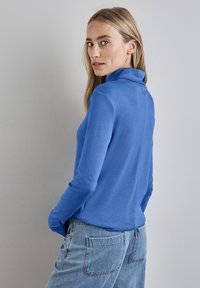 Blå långärmad topp med hög krage, tillverkad av slät tyg. Har en smal passform och detalj med söm bak, kombinerad med ljusblå jeans.