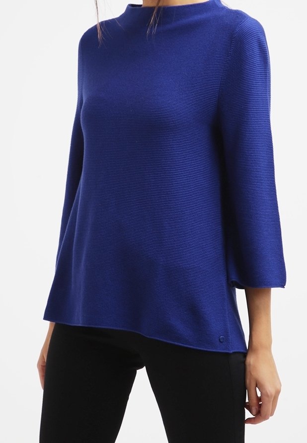 Femme portant un pull en maille côtelée bleu à manches trois-quarts et un pantalon noir, posant devant un fond blanc.
