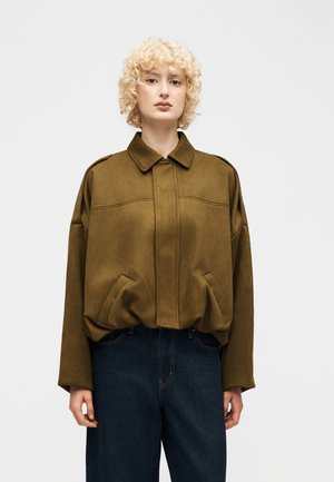 Mladá osoba s kučeravými blond vlasmi nosí nadrozmernú olivovo zelenú bundu s veľkými vreckami a tmavomodré rifle, stojí na bielom pozadí.