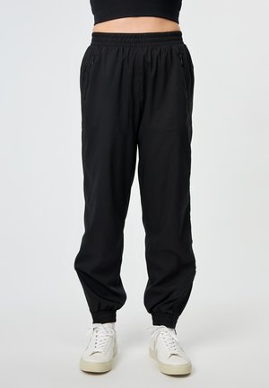 Person wearing black elastic-waist jogger pants with side zip pockets and white sneakers against white background.
