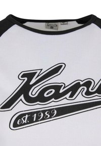 Bílé tričko s krátkým rukávem s černými raglánovými rukávy. Na přední straně se nachází velké černé logo "Kanu" a text "est. 1989" pod ním.