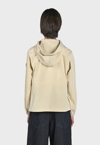 Giacca beige con cappuccio, polsini elasticizzati, texture liscia e design aerodinamico. La vista posteriore mette in evidenza uno stile semplice e minimalista.