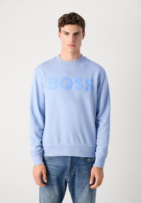 Sudadera azul claro con cuello redondo y puños de canalé, con un gran texto "BOSS" en un color a juego en la parte frontal. Jeans de mezclilla.