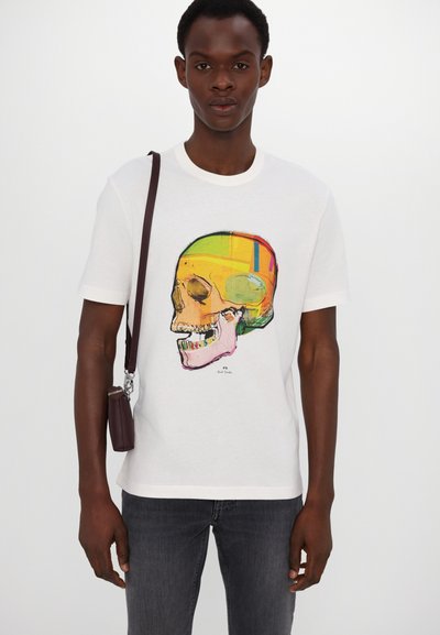 Camiseta de algodón blanca con un gráfico de calavera colorido, con contornos negros y acentos en amarillo, rosa y verde. Llevada con pantalones vaqueros oscuros.