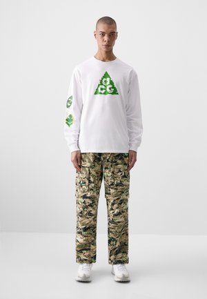 White long-sleeve shirt with green triangle logo and leaf graphics on sleeves; camo cargo pants with hues of green and tan; white sneakers.