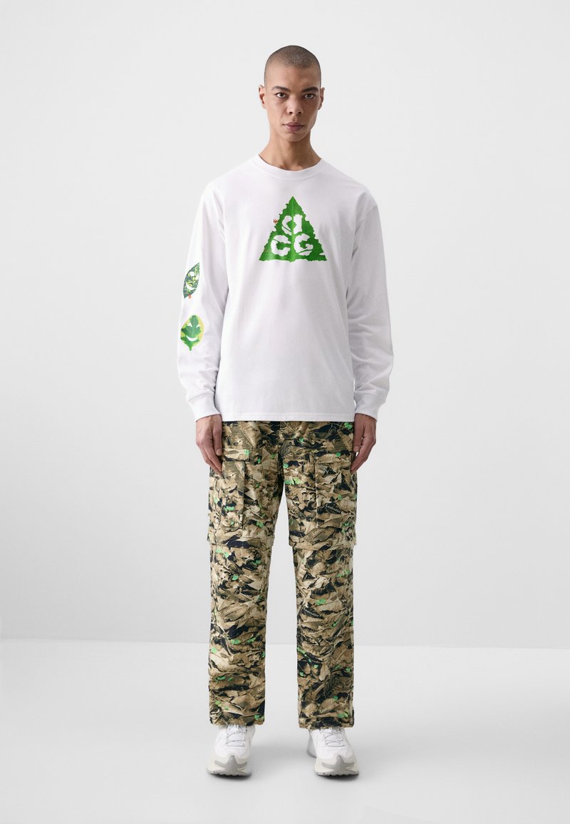White long-sleeve shirt with green triangle logo and leaf graphics on sleeves; camo cargo pants with hues of green and tan; white sneakers.