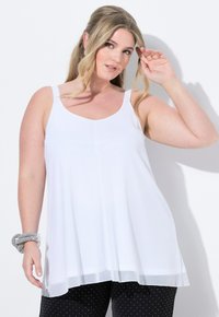 Top bianco senza maniche con scollatura a goccia, caratterizzato da un design fluido e un orlo trasparente. Abbinato a pantaloni neri a pois e un bracciale argentato.