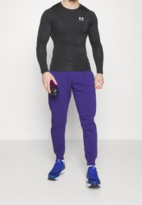 Svart långärmad träningsskjorta, lila joggerbyxor och blå löparskor; håller en svart vattenflaska, med logotyp på skjortan.