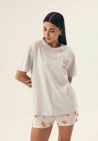 Biała bawełniana koszulka z krótkim rękawem z napisem "C'est la Vie" w złotym kolorze. W zestawie z białymi szortami ozdobionymi pomarańczowymi wzorami.