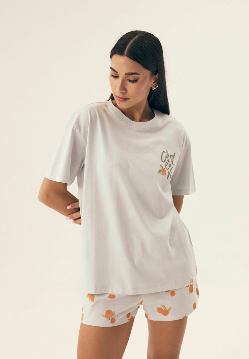 Biała bawełniana koszulka z krótkim rękawem z napisem "C'est la Vie" w złotym kolorze. W zestawie z białymi szortami ozdobionymi pomarańczowymi wzorami.