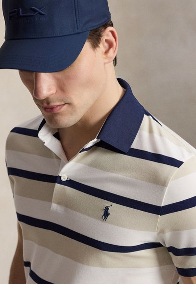 Homme portant une casquette bleu marine et un polo rayé beige, blanc et bleu marine avec un col bleu marine et un petit logo brodé sur la poitrine.