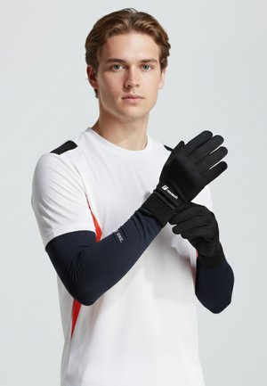 Young man wearing white and black athletic wear puts on black gloves with logo on a plain light background.