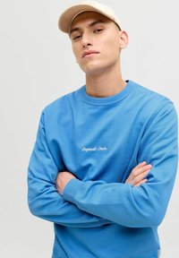 Mėlynas megztinis su apvaliu iškirpimu, ribotu rankogaliu ir siuvinėtus "Originals Studio" tekstu ant krūtinės. Dėvimas su béžo spalvos kepure. Lycra textūra.