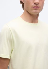 Νεαρός άνδρας που φοράει κοντομάνικη μπλούζα Puma ανοιχτού κίτρινου χρώματος με ένα διακριτικό λευκό λογότυπο γάτας που πηδάει στο στήθος, σε απλό φόντο.