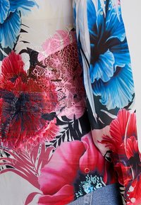 Blouse fluide avec un motif floral dans des teintes vif de bleu, rouge et rose, en tissu transparent avec des détails en dentelle.