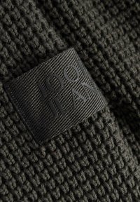 Svart stickad textil med en framträdande rektangulär svart etikett, präglad med "JOOP JEANS" i ett elegant, modernt teckensnitt. Slät textur.