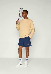 Beige sweatshirt og marineblå shorts med folder, hvit logo. Hvite sneakers med blå og hvite stripete sokker, holder en tennisracket.