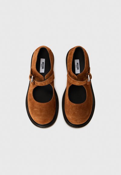 Brown suede shoes with open sides, rounded toes, and a buckled strap across the upper. Black soles contrast with the soft texture.