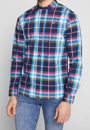Mann trägt ein langärmliges Hemd mit Karo-Muster in Marineblau, Pink, Blau, Weiß und Oliv, kombiniert mit blauen Jeans.