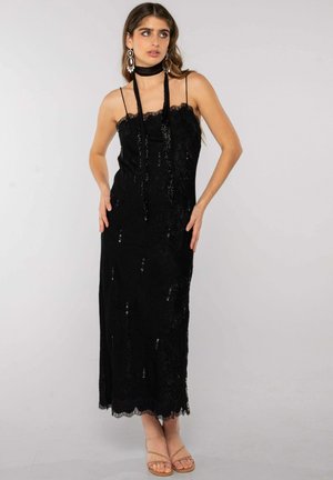 Mujer con un vestido largo negro de encaje con tirantes finos, una bufanda negra a juego para el cuello, pendientes grandes y sandalias beige abiertas en la punta.