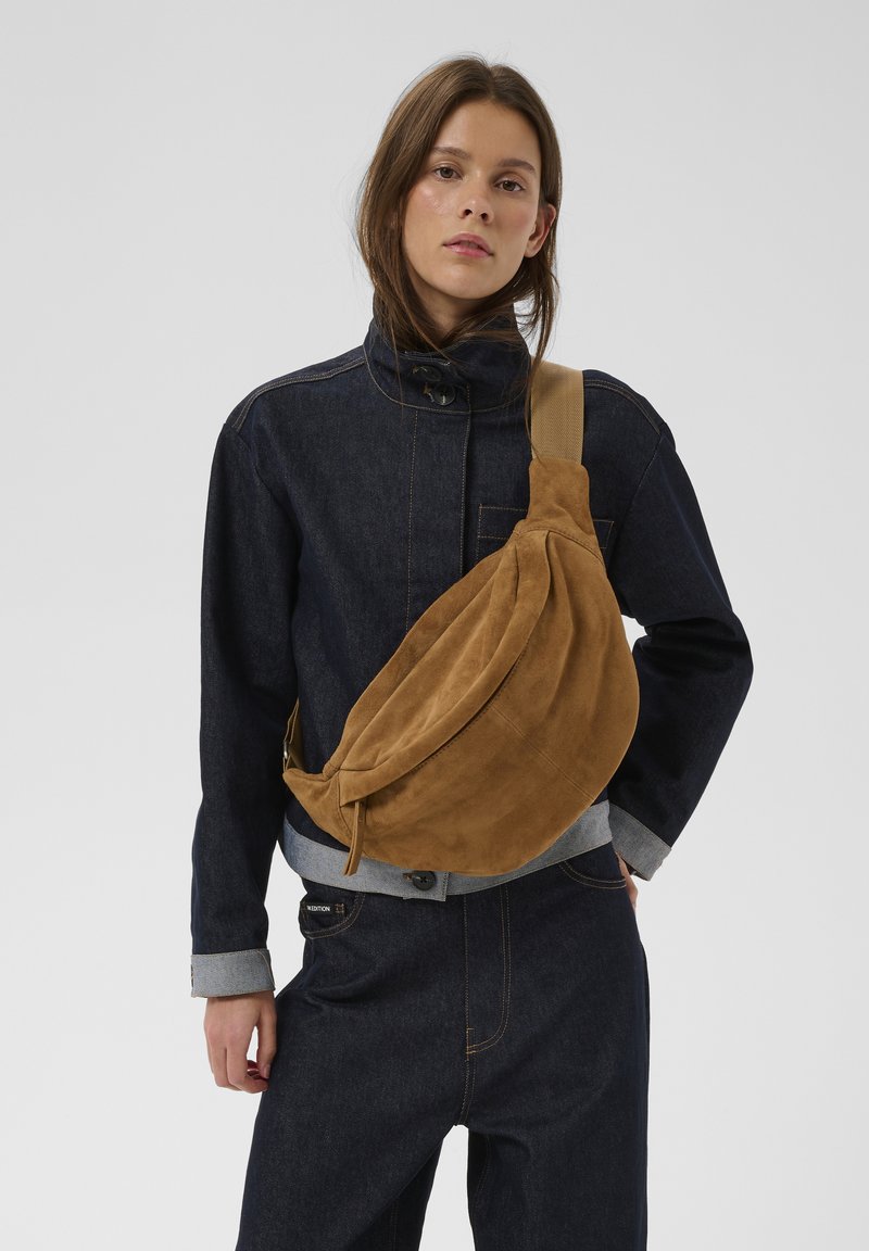 Jonge vrouw met een donker denim jack en broek, met een grote bruine suède crossbodytas voor een effen grijze achtergrond.