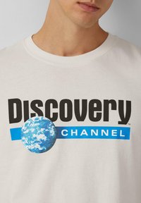 Jasno-beżowy t-shirt bawełniany z odważnym czarnym napisem "DISCOVERY" i "CHANNEL", z niebieską grafiką Ziemi na niebieskim pasku. Okrągły dekolt.