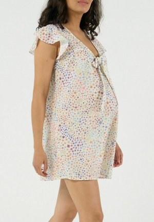 Vrouw draagt een korte, veelkleurige bloemenjurk met fladdermouwen en een strik aan de voorkant, staand tegen een effen witte achtergrond.