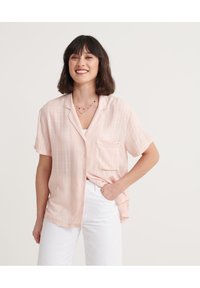Blusa rosa chiaro a maniche corte con colletto a polo, tasca frontale e texture sottile. Abbinata a pantaloni bianchi.