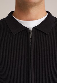 Zwarte geribbelde rits kleding met een gekraagde hals, voorzien van een kleine rits en een witte laag shirt eronder. Textuur van de stof is zichtbaar.