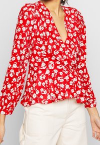 Röd blus med blommönster, långa ärmar och djup V-ringning. Har vita blommönster och en rynkad nederkant för en peplum-effekt.